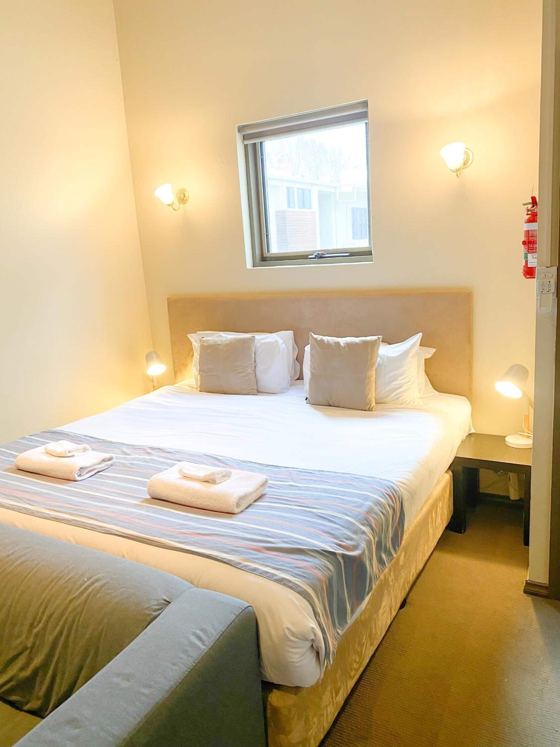 A hotel room with a large bed, two bedside lamps, a window, and a fire extinguisher on the wall.
