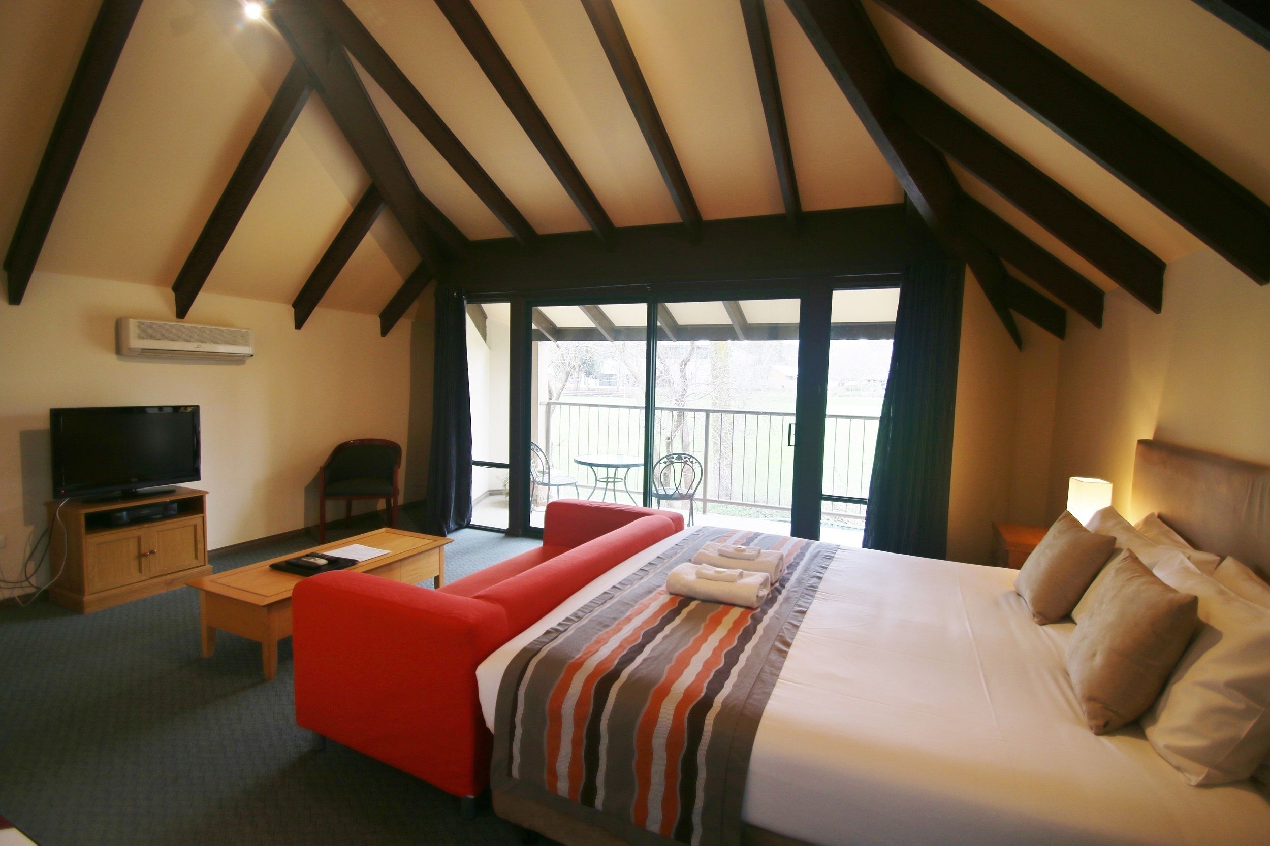 Habitación de hotel con cama, televisión, sofá rojo, mesa de madera y balcón con vistas.