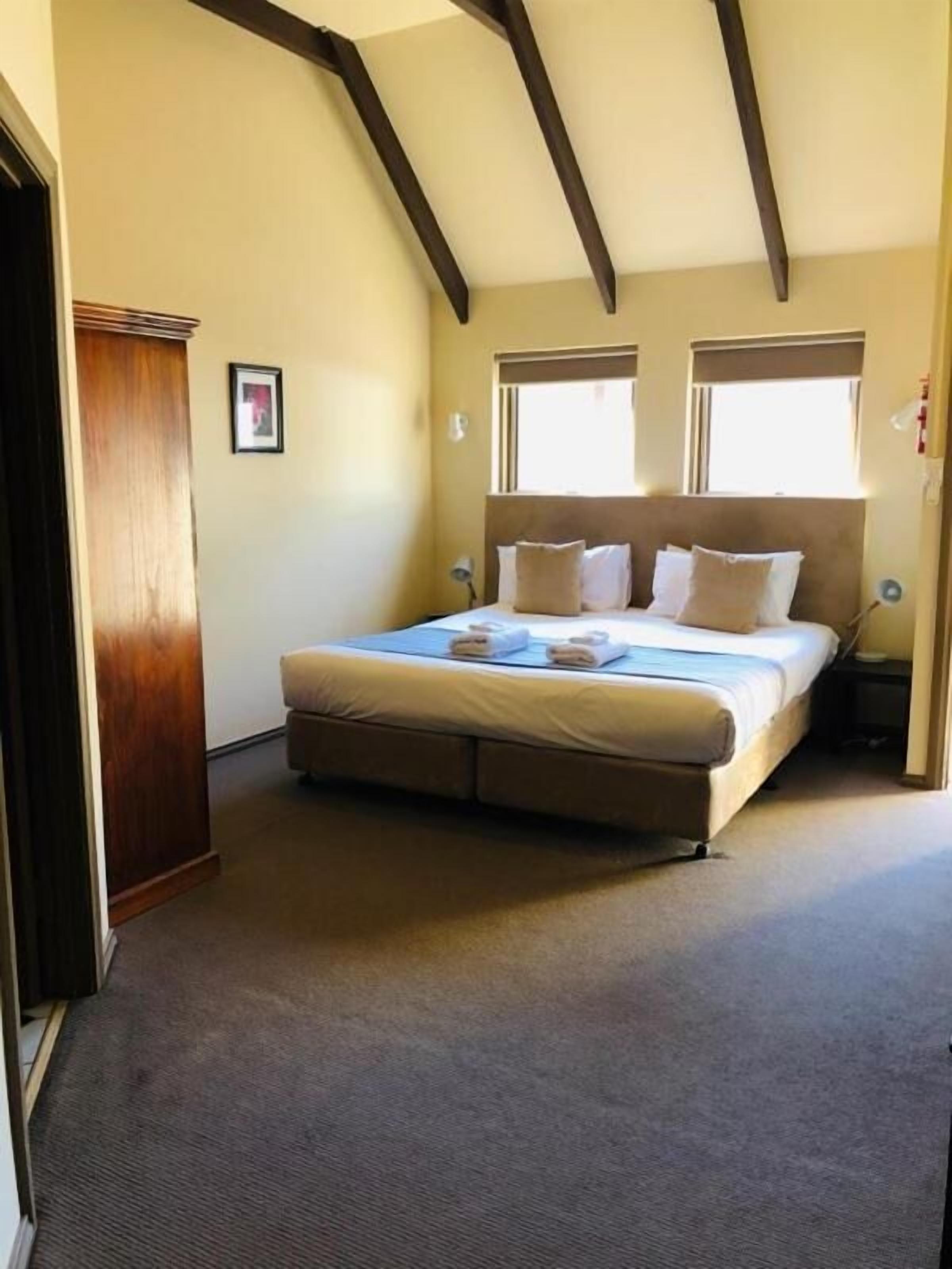A bedroom with a large bed, wooden beams on the ceiling, two windows, and a door.