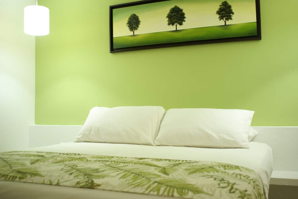 Un lit avec une literie blanche et un couvre-lit à motifs de feuilles vertes, un mur blanc et un tableau encadré représentant des arbres accroché au mur.