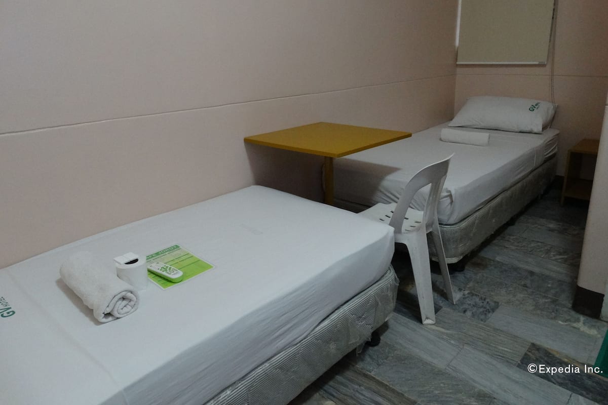 Quarto de hotel pequeno com duas camas de solteiro, uma mesa, uma cadeira e um sanitário.
