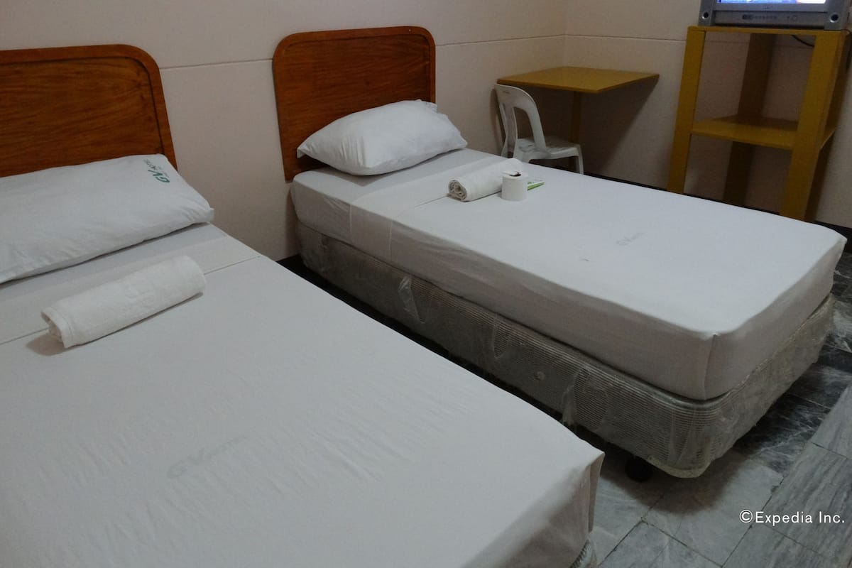 Quarto de hotel com duas camas de solteiro, uma mesinha de madeira, uma televisão e uma cadeira branca.