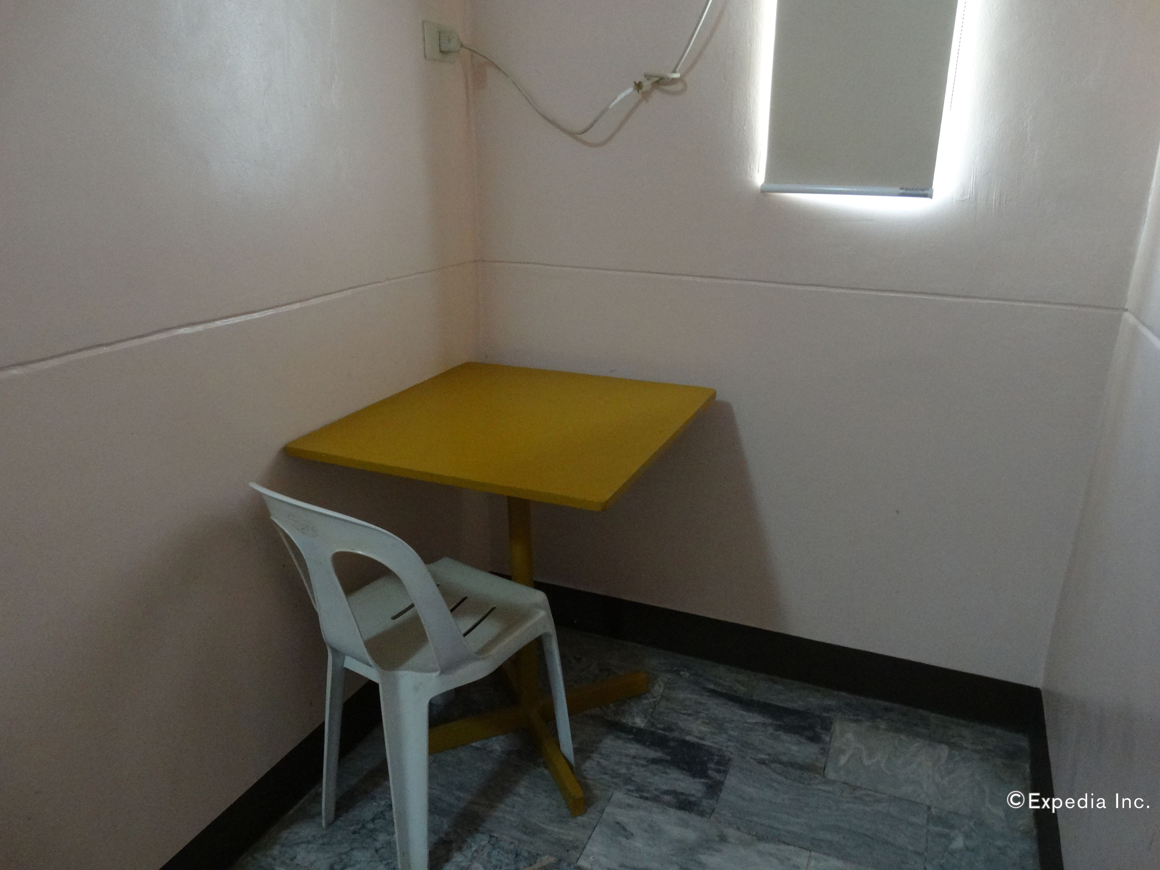 Una habitación pequeña con una mesa amarilla y una silla de plástico blanca. Se ve una ventana con vistas al exterior.