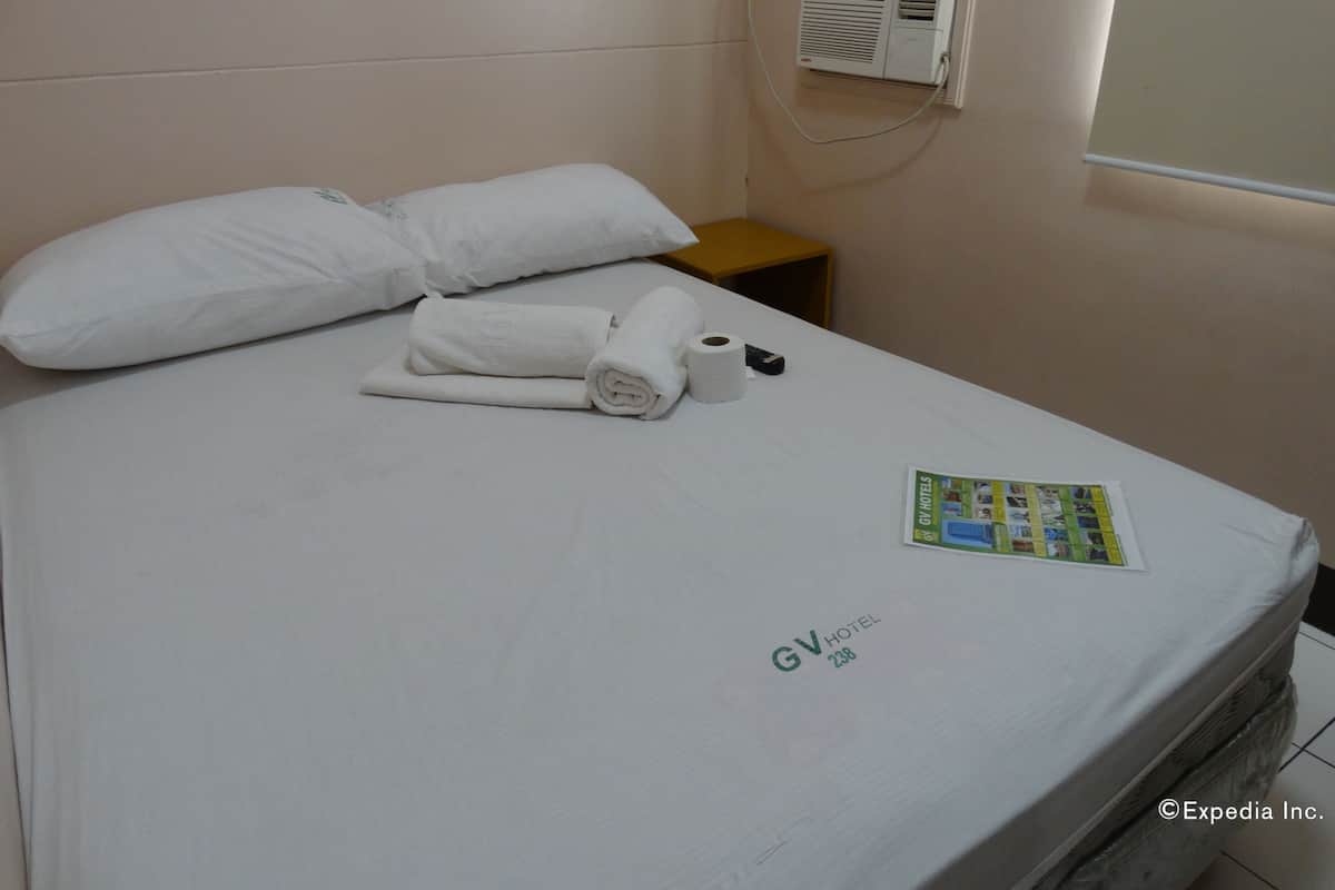 Quarto de hotel com uma cama, travesseiros, um controle remoto, um rolo de papel higiênico e um folheto sobre a cama.
