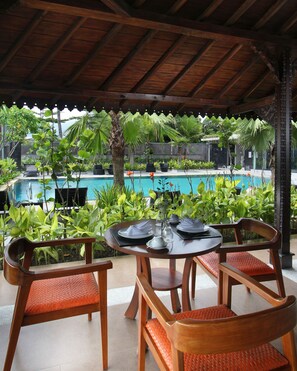 A covered outdoor seating area with a pool view, featuring wooden chairs and a table set for dining.