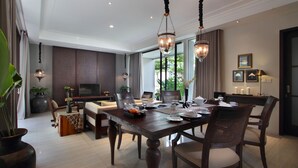 A dining area with a wooden table set for four, surrounded by chairs. There is a television and a sofa in the background, and the room has large windows with curtains.