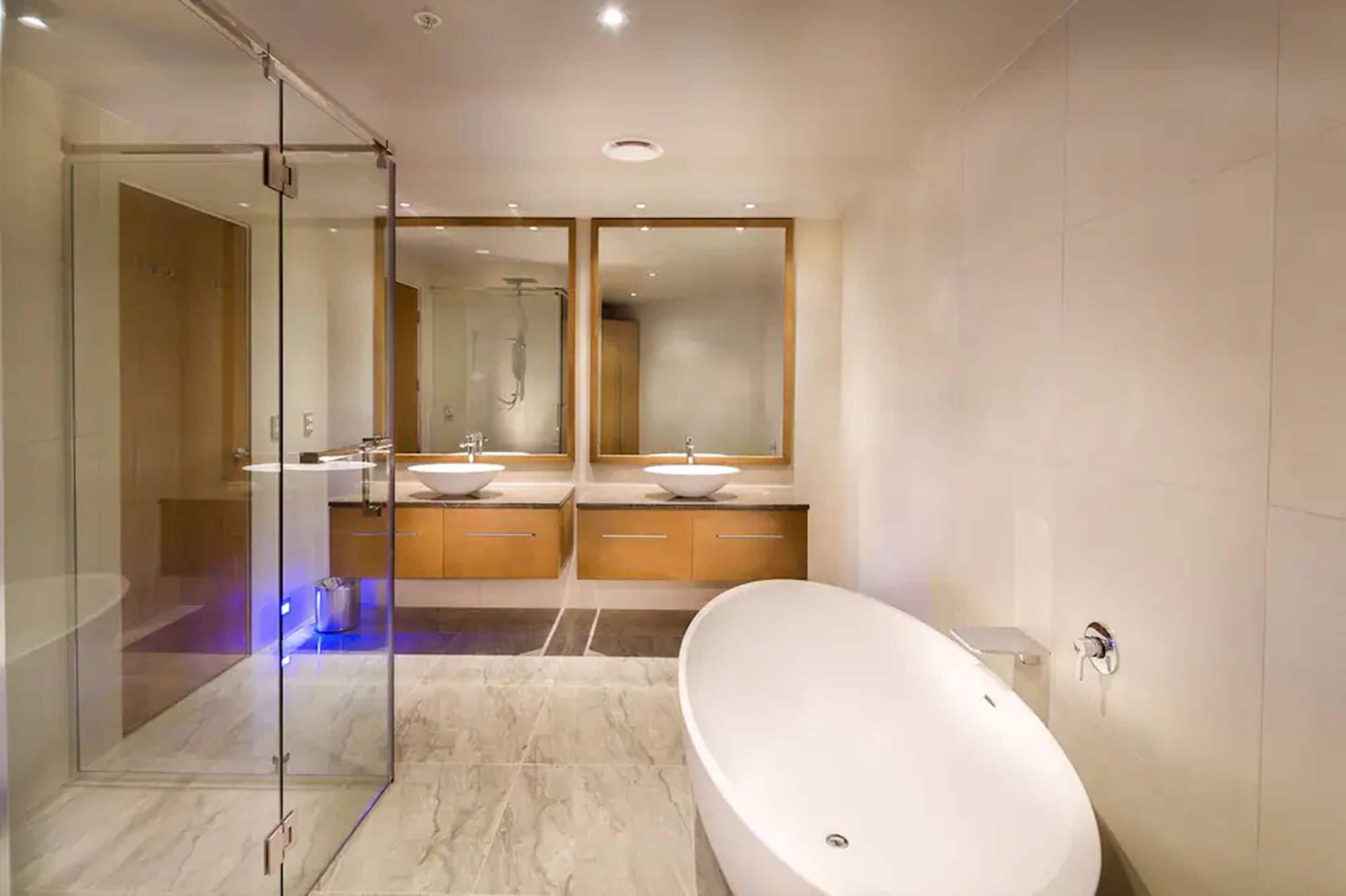 Un baño moderno con cabina de ducha de vidrio, dos lavamanos con espejos y una bañera independiente.