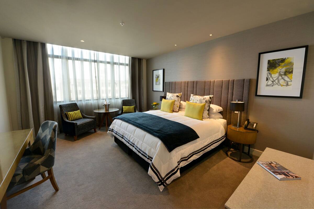 A modern hotel room with a large bed, a desk, a chair, and a view of the city through large windows.