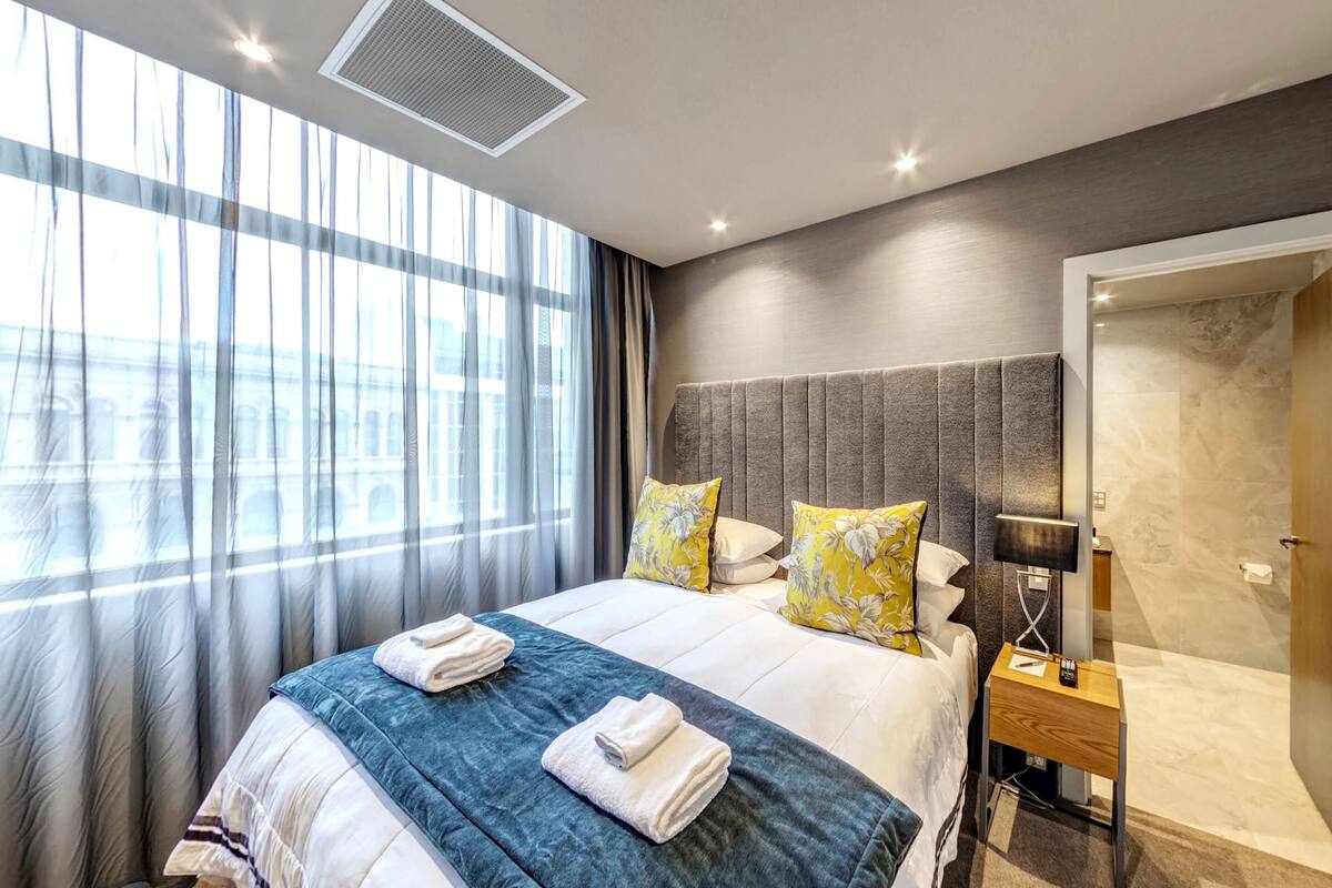A hotel room with a large bed, a bedside table with a lamp, a wardrobe, and a view of the city through the window.