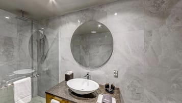 Un baño moderno con ducha de vidrio, un espejo ovalado grande y un tocador con loseta de mármol, un lavamanos y un perchero para toallas.