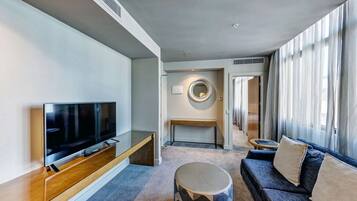Una habitación de hotel moderna con televisión de pantalla plana, un sofá, una mesita de centro redonda y un amplio ventanal con cortinas traslúcidas.