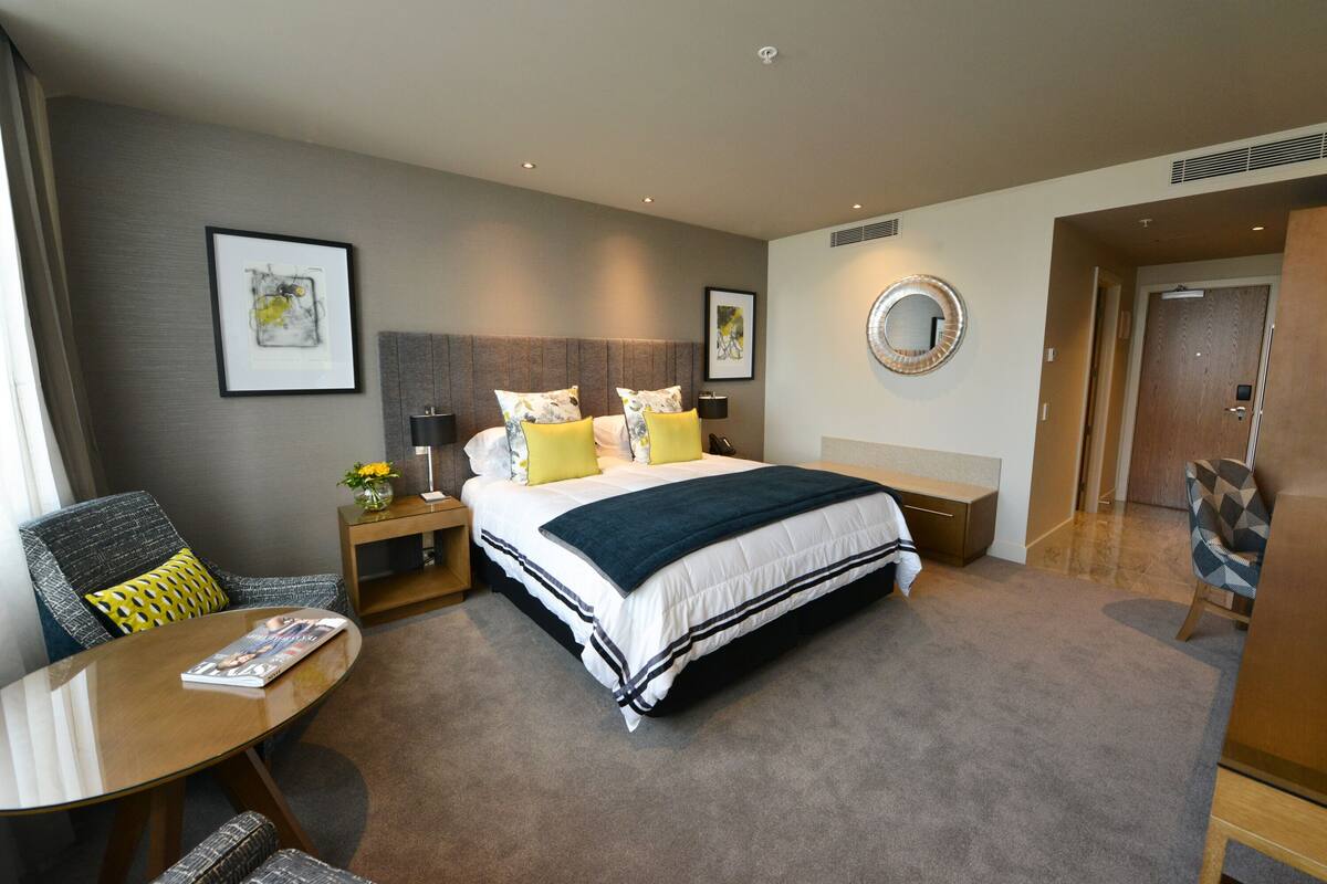 A hotel room with a large bed, a bedside table with a lamp, a chair, a round table, and artwork on the walls.