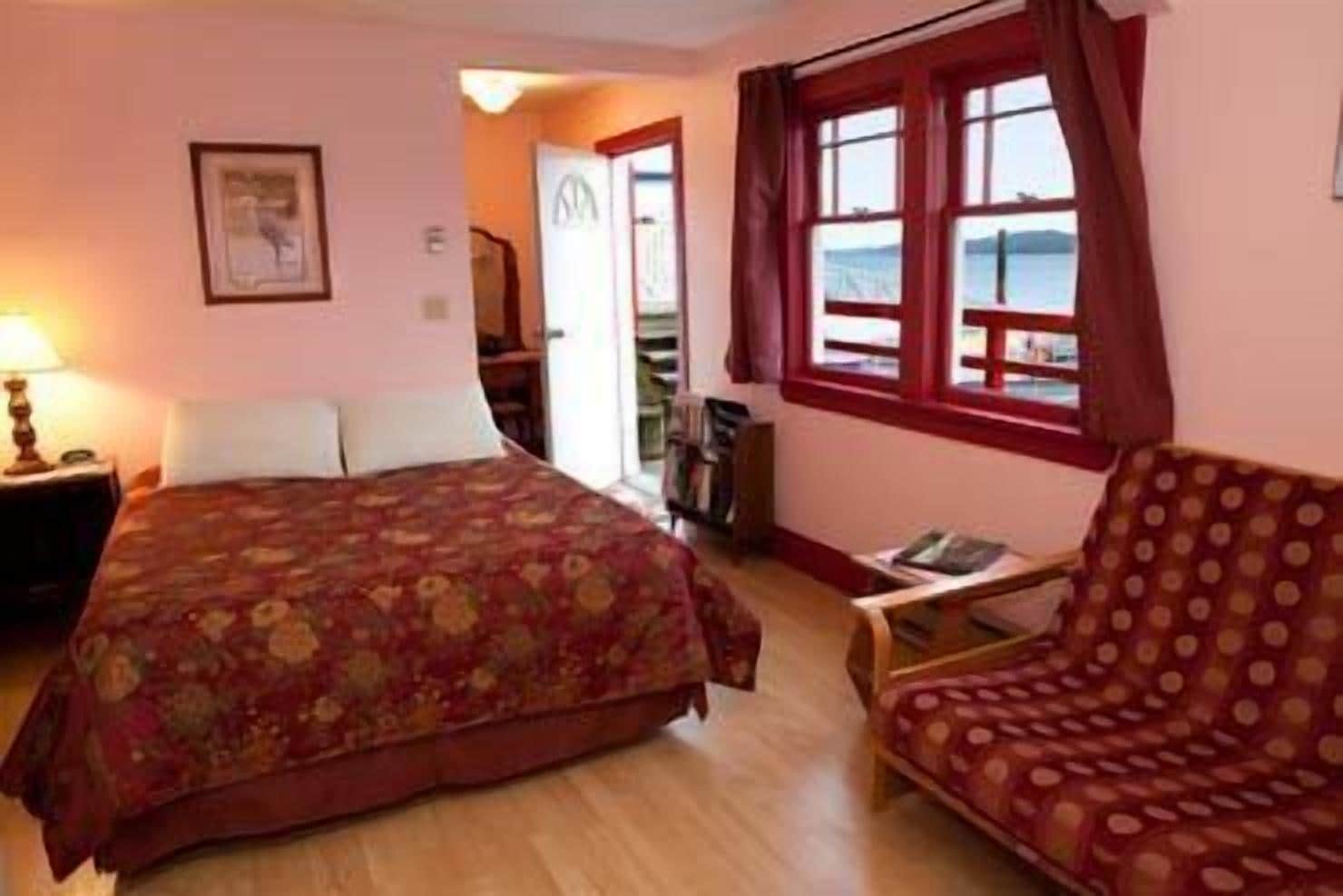 A bedroom with a bed, a chair, a nightstand, a window with curtains, and a door leading to another room.
