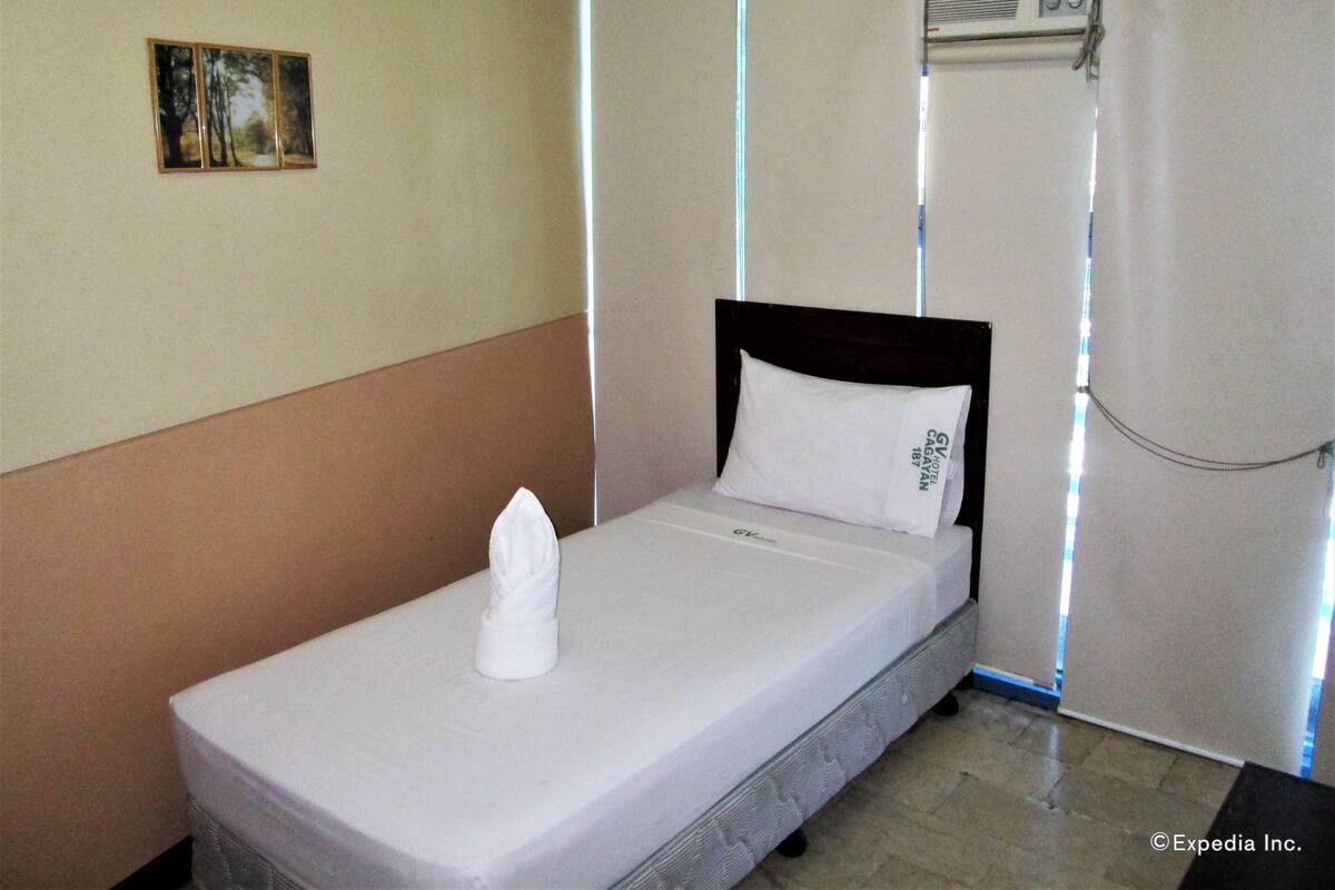 Una cama individual con colchón y almohada blancos, cabecera, un cuadro pequeño en la pared y un equipo de aire acondicionado sobre la cama.
