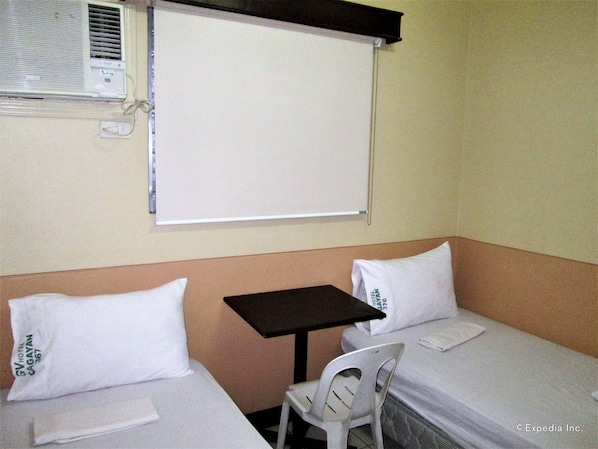 Una habitación de hotel pequeña con dos camas, una mesa, una silla, una ventana con persiana y un equipo de aire acondicionado.