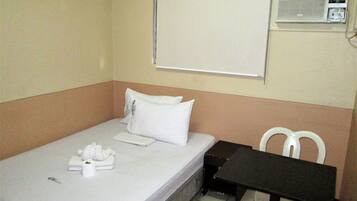 Une petite chambre avec un lit simple, recouverte d’une courtepointe blanche, un oreiller et une serviette pliée. Une petite table et une chaise se trouvent à droite du lit. Un appareil de climatisation est fixé au mur, au-dessus du lit.