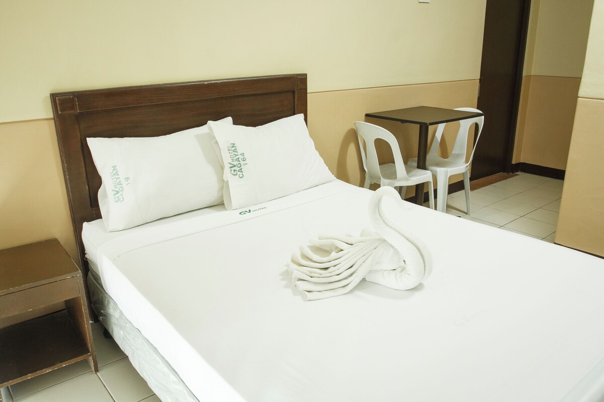 Una cama bien tendida con sábanas y almohadas blancas, una toalla blanca doblada y dos sillas de plástico blanco junto a una pequeña mesa.