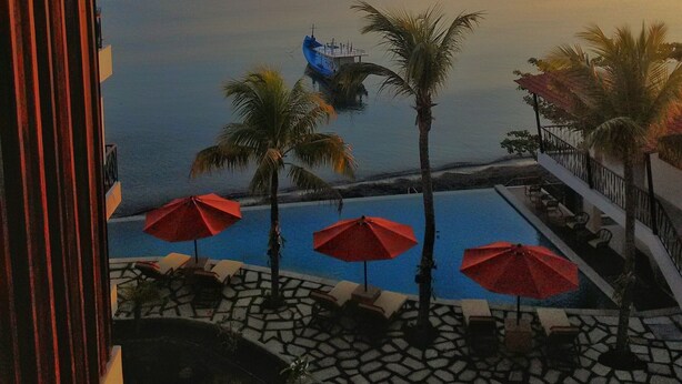 A resort view with a pool area, palm trees, and a boat on the water. - Capa Maumere (Maumere)
