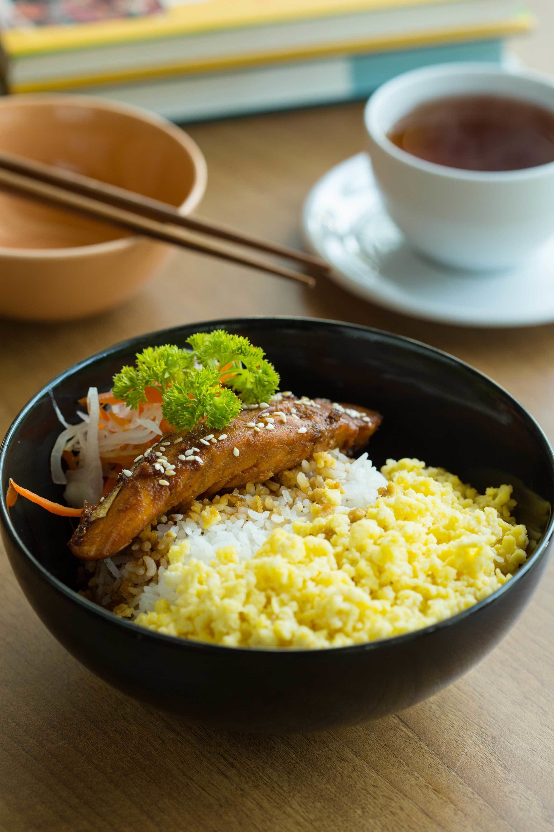 Un tazón de arroz con un trozo de carne y guarniciones sobre una mesa.