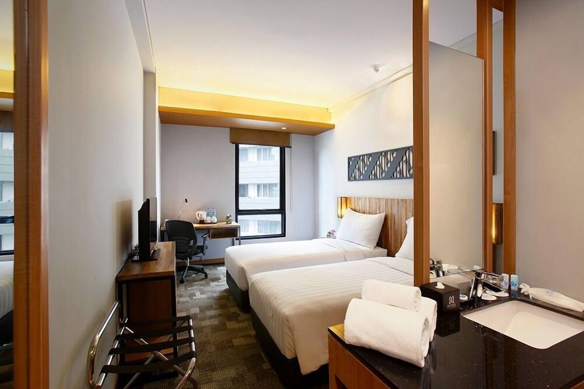 A hotel room with two beds, a desk with a computer, a chair, a desk lamp, a mirror, and a bathroom with a sink and towel.