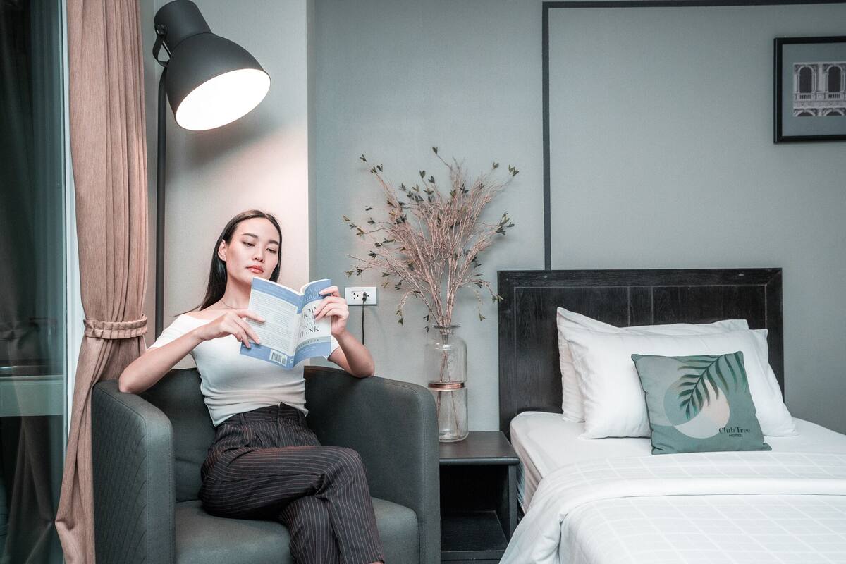 A woman reading a book in a modern hotel room with a bed, a nightstand, and a vase with branches.