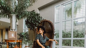 A woman seated in a wicker chair in a spacious room with large windows, a checkered floor, and various potted plants. - Club Tree Hotel (Songkhla)