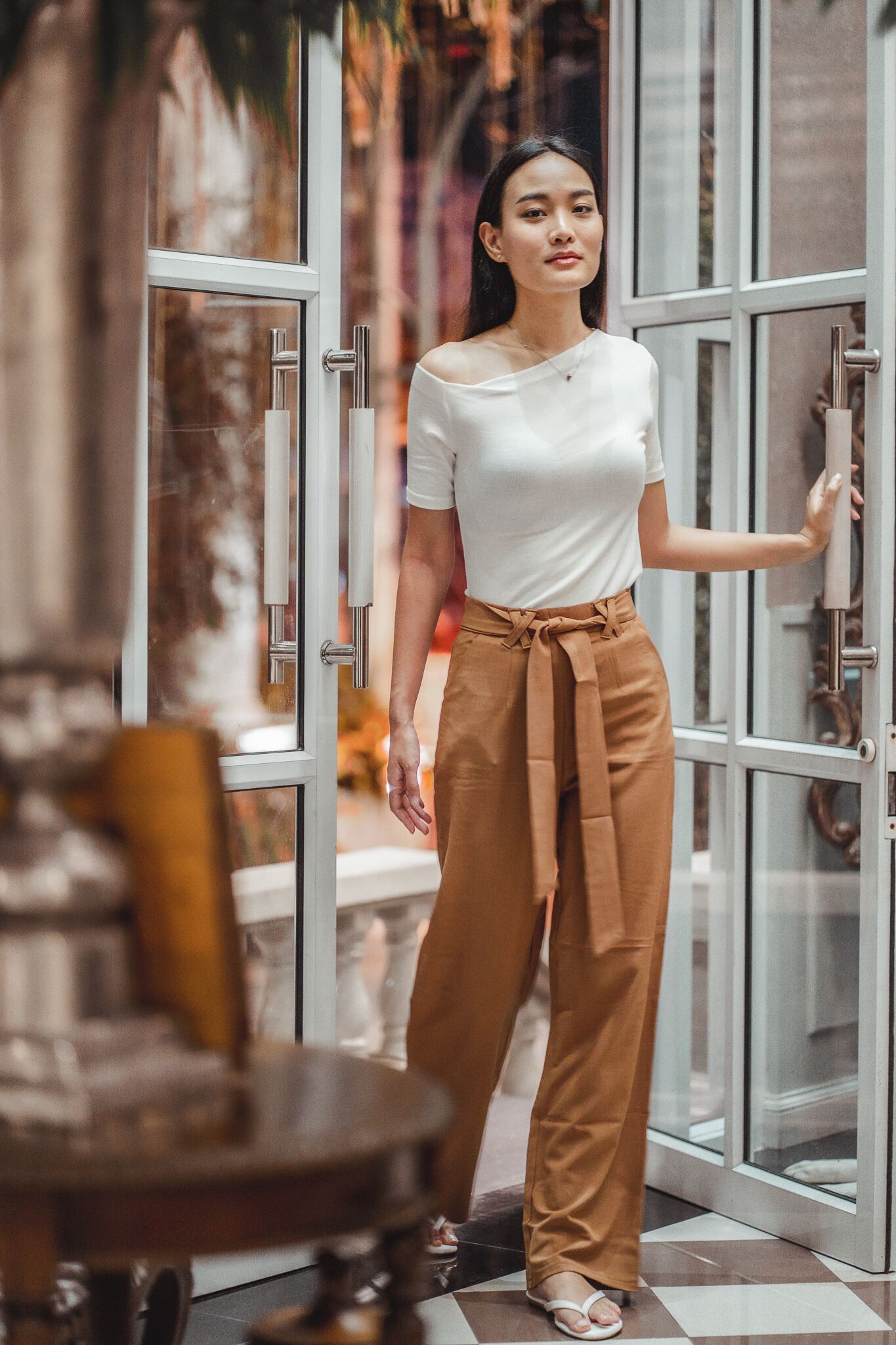 A person standing in front of a glass door with a metal handle, wearing a white top and brown pants.