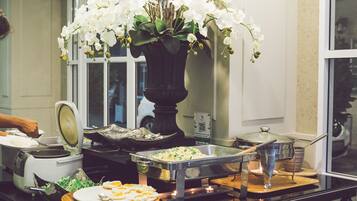 A buffet setup with various food items, a large vase with white flowers, and a window in the background.