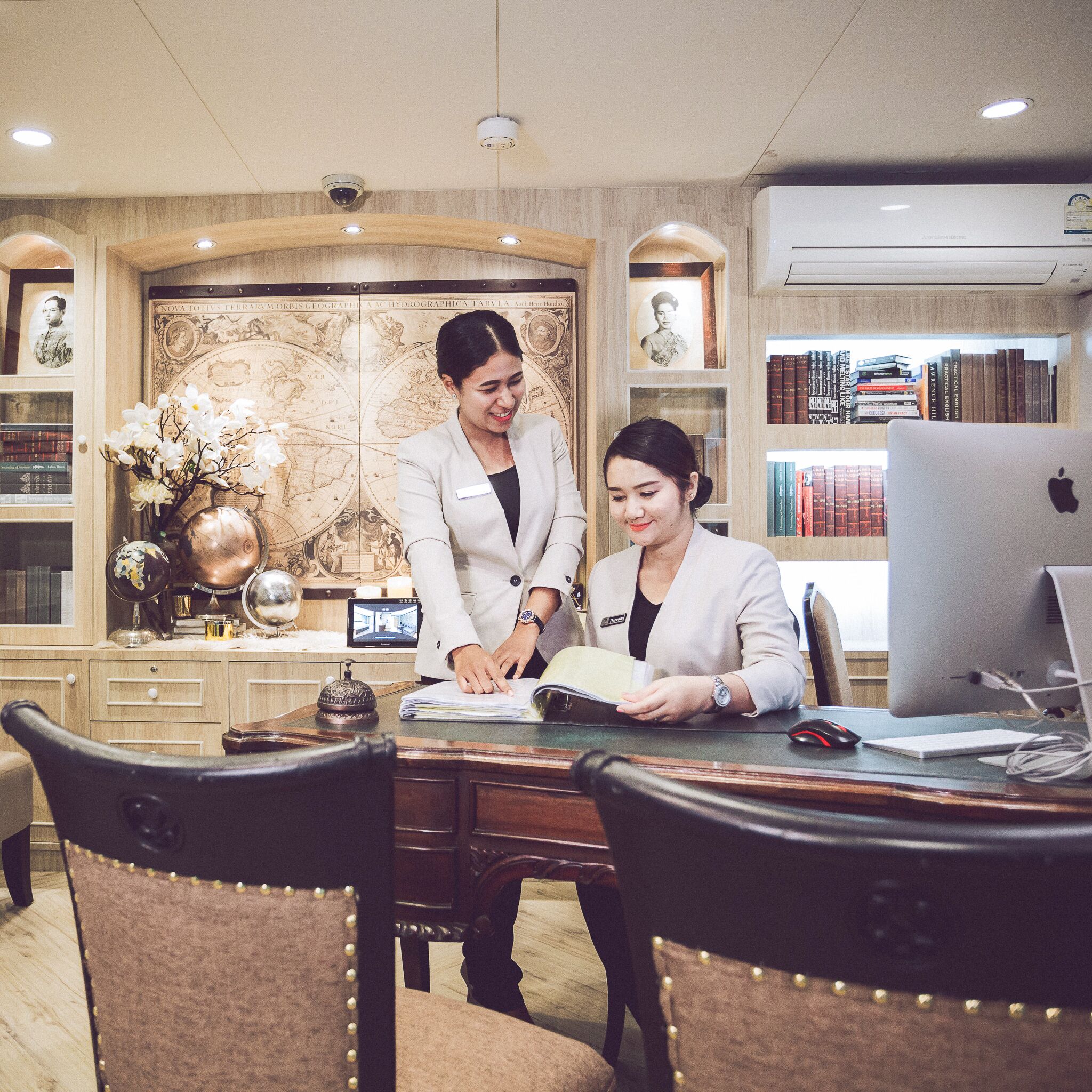 A professional office setting with two individuals at a desk, surrounded by bookshelves, a globe, and a decorative world map.