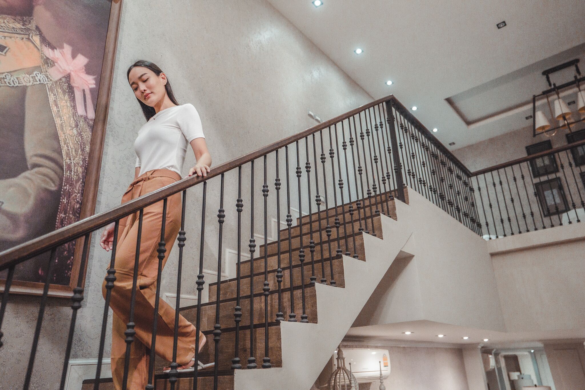 A woman descending a staircase with a metal railing, a large framed artwork on the wall, and a modern interior design.