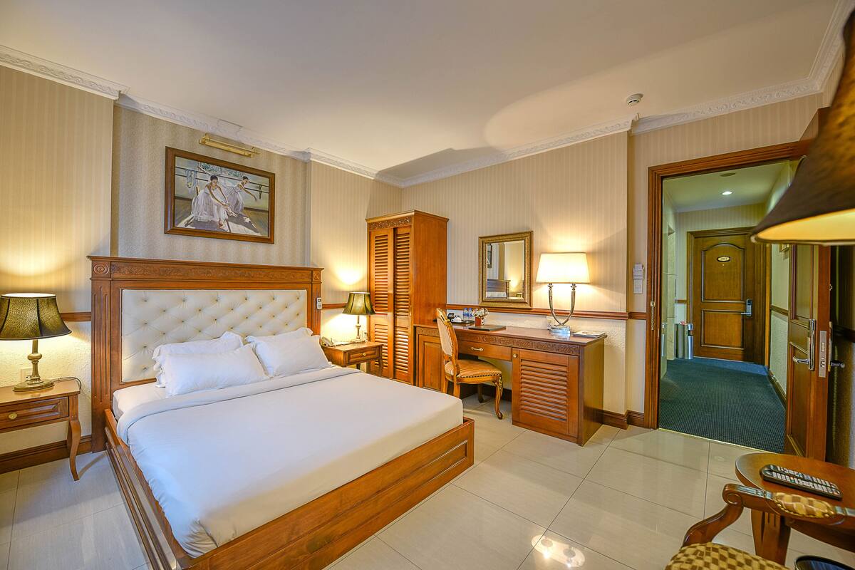 Une chambre d’hôtel avec un grand lit, un bureau, une chaise, un miroir et une porte donnant sur une autre pièce.