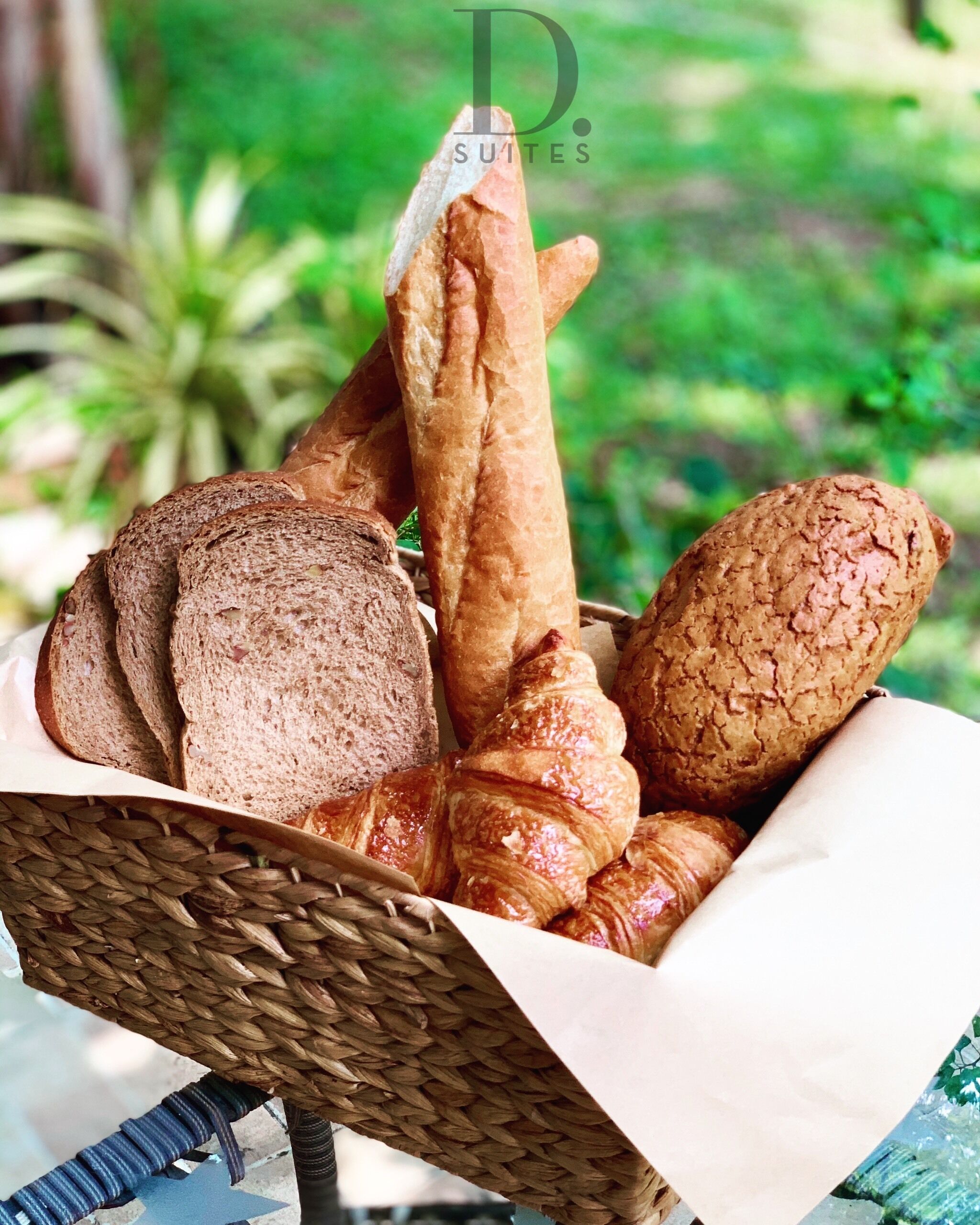 A basket of baguettes and croissants on a table with a green outdoor background.