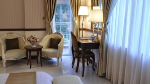 Una habitación con dos sillones, una mesita con un jarrón de flores, un escritorio con una lámpara y vistas a través de una ventana.