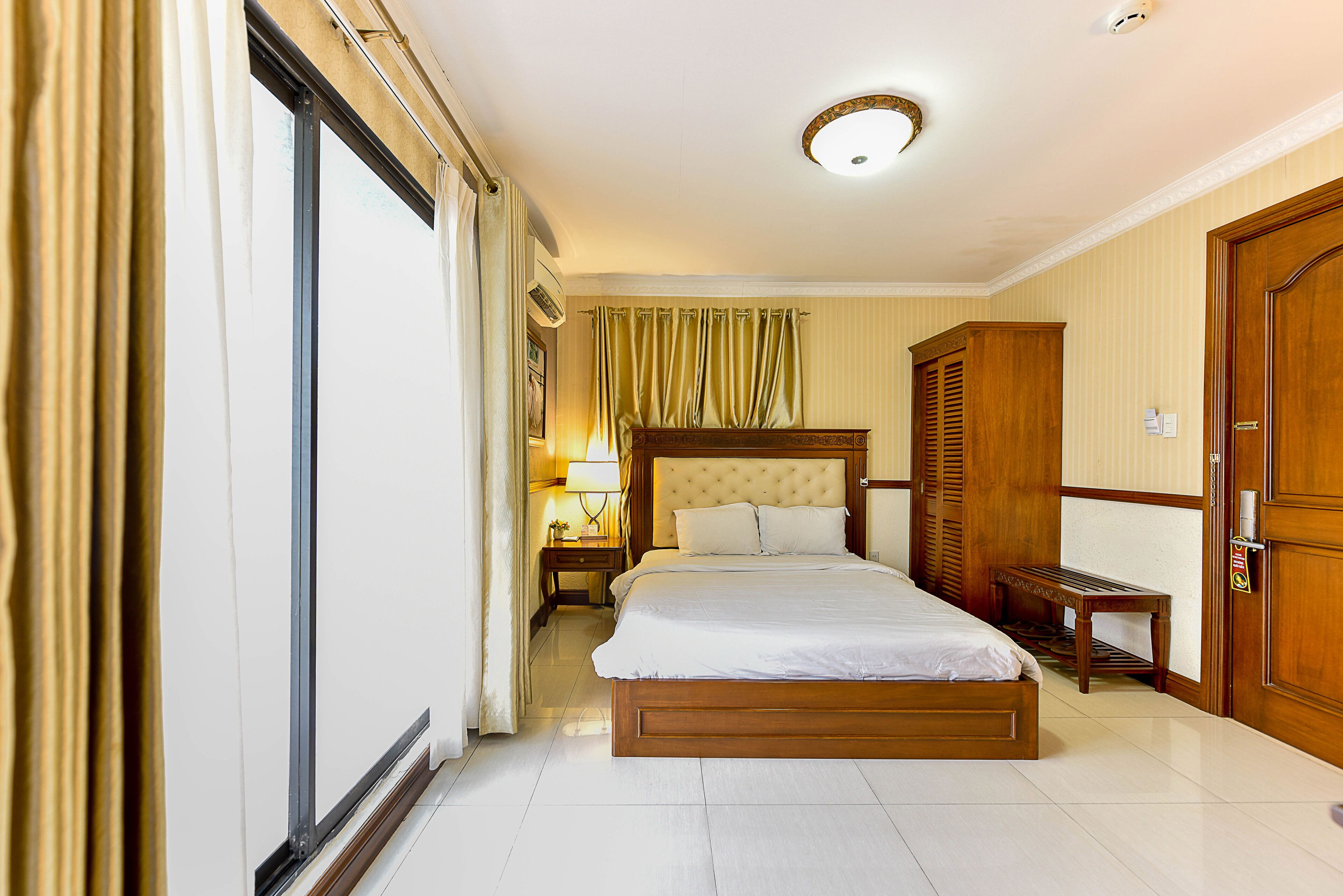 Een hotelkamer met een groot bed, een houten garderobe, een nachtkastje met een lamp en een bankje bij het raam.