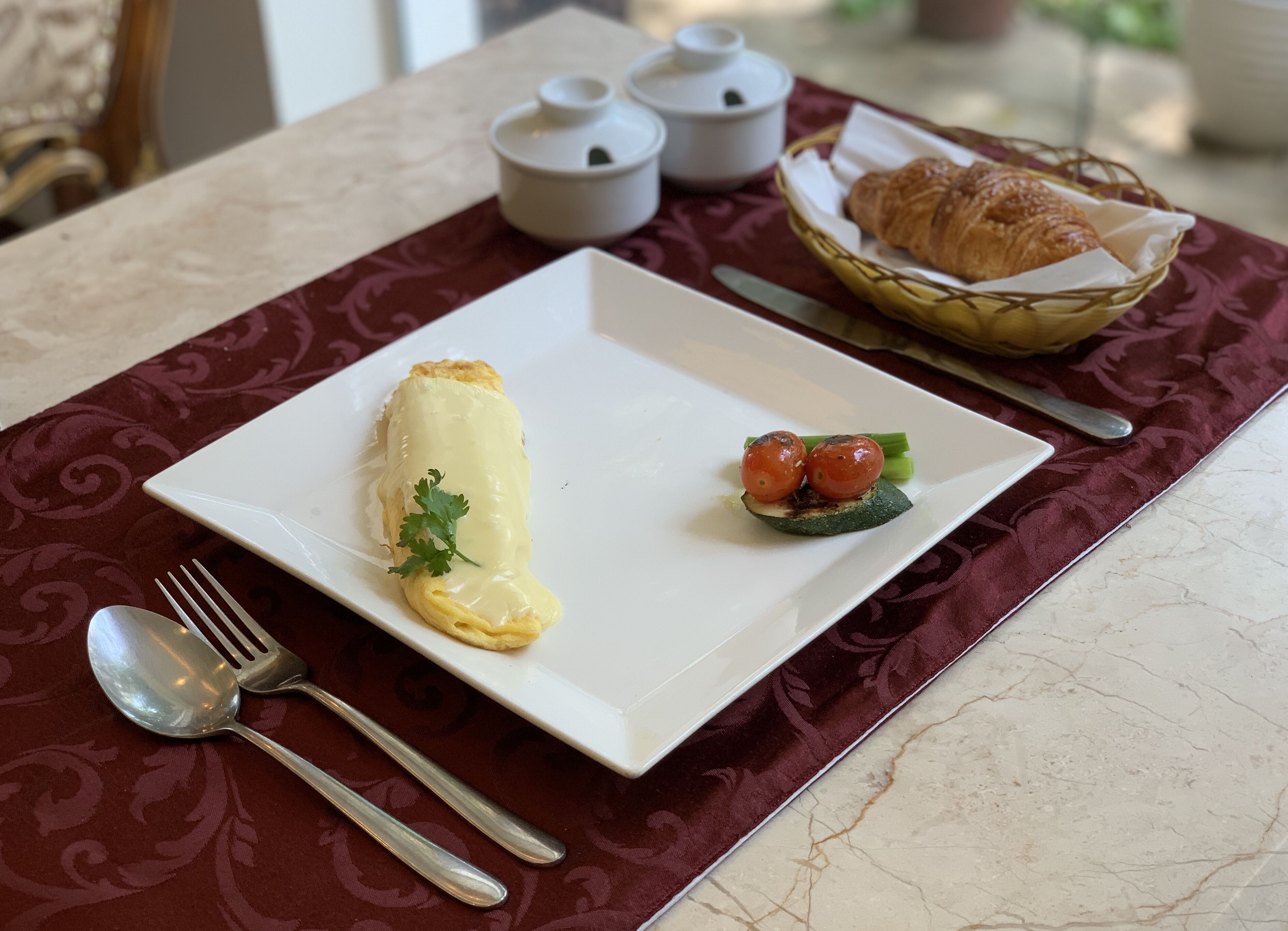 A breakfast plate with an omelette, cherry tomatoes, and a croissant on a marble table with a red placemat.