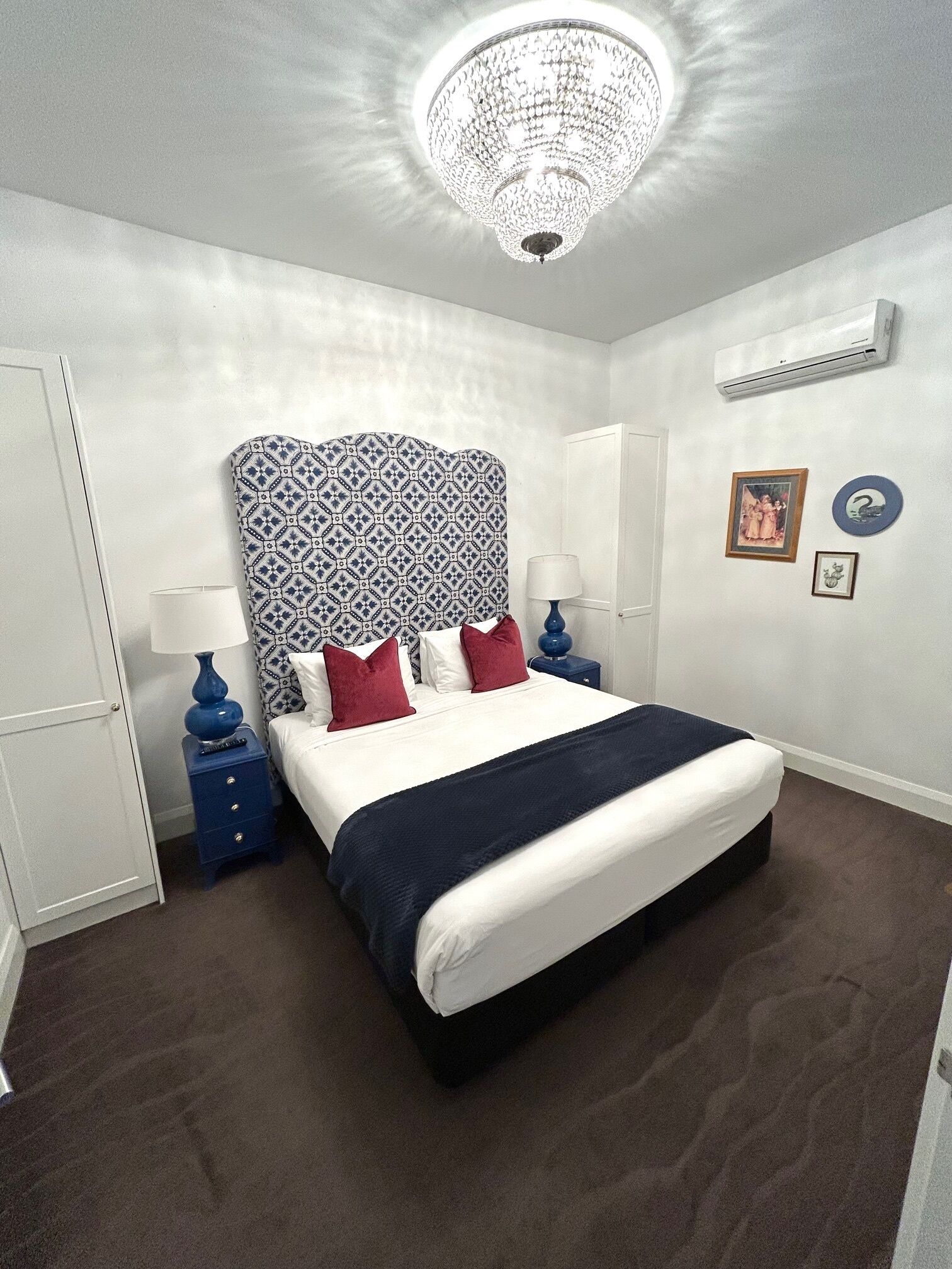 A neatly arranged bedroom with a large bed, a patterned headboard, a chandelier, a bedside table with a lamp, and a wall-mounted air conditioner.