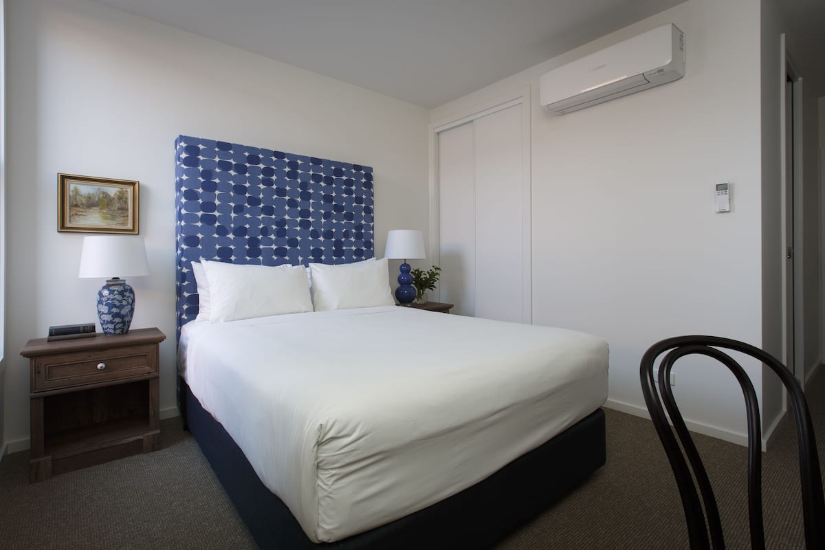 Una cama bien hecha con cabecera azul, dos mesitas de noche con lámparas, un cuadro enmarcado en la pared y un equipo de aire acondicionado.