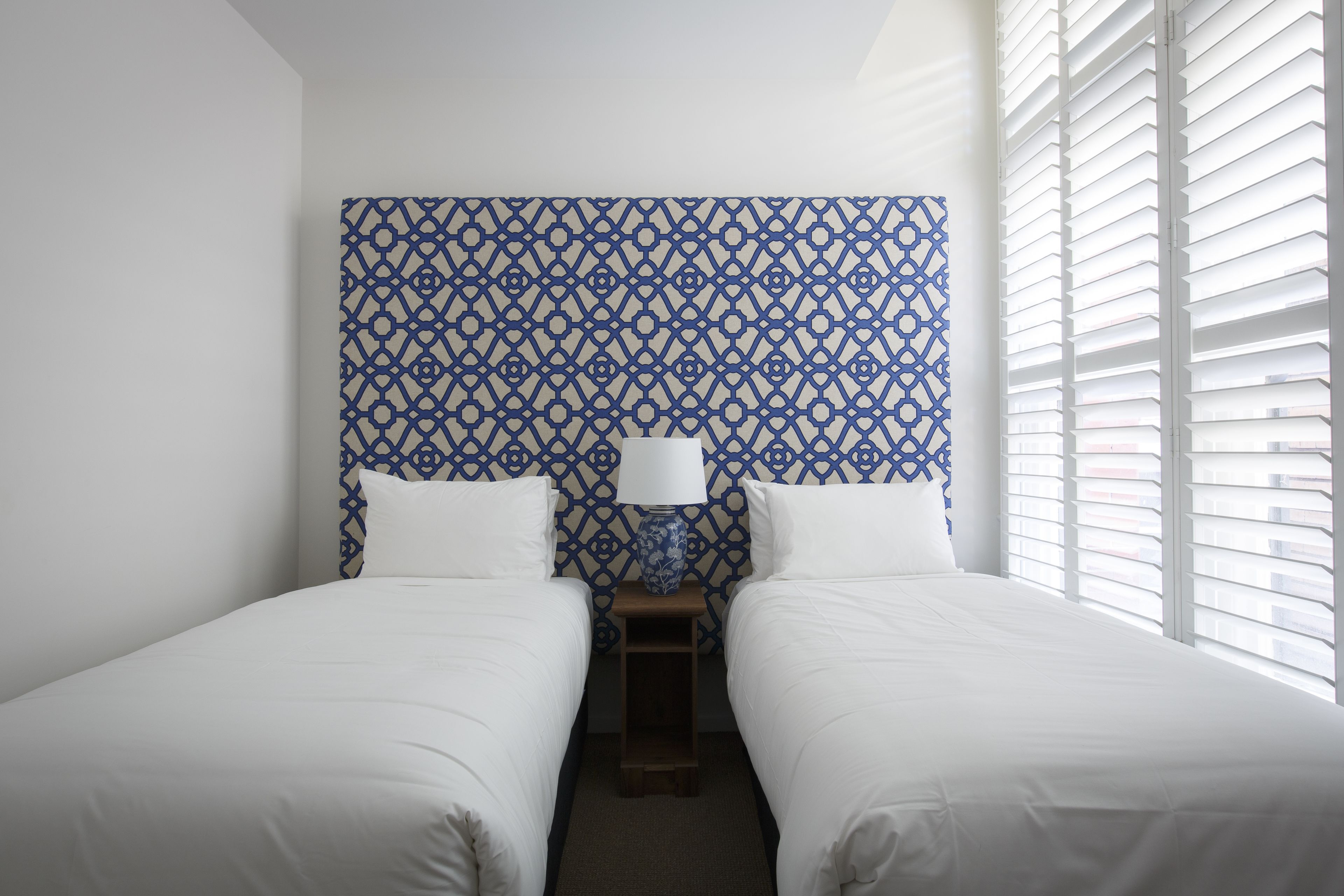 Une chambre d’hôtel avec deux lits, une tête de lit à motifs, une table de chevet avec une lampe et une fenêtre avec des stores.