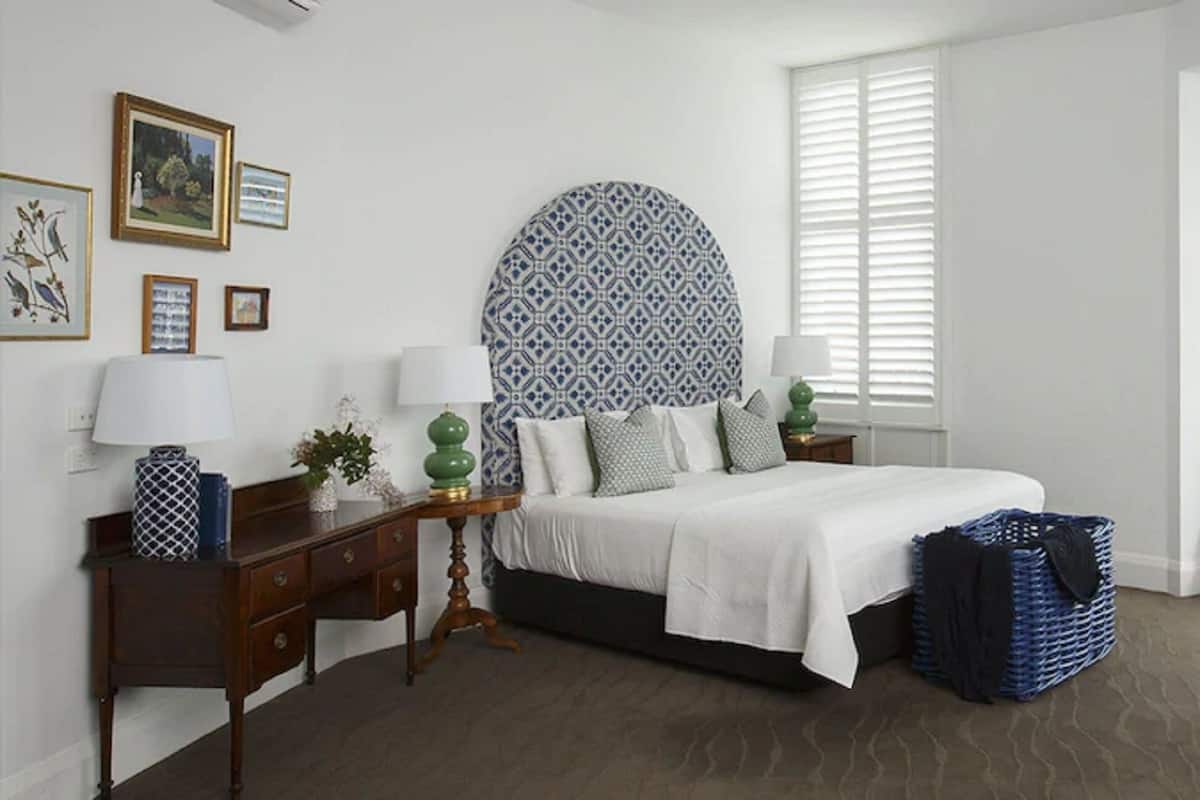 Un dormitorio con cama, mesitas de noche, cómoda, silla, ventana con persianas y un gran elemento decorativo en la pared.