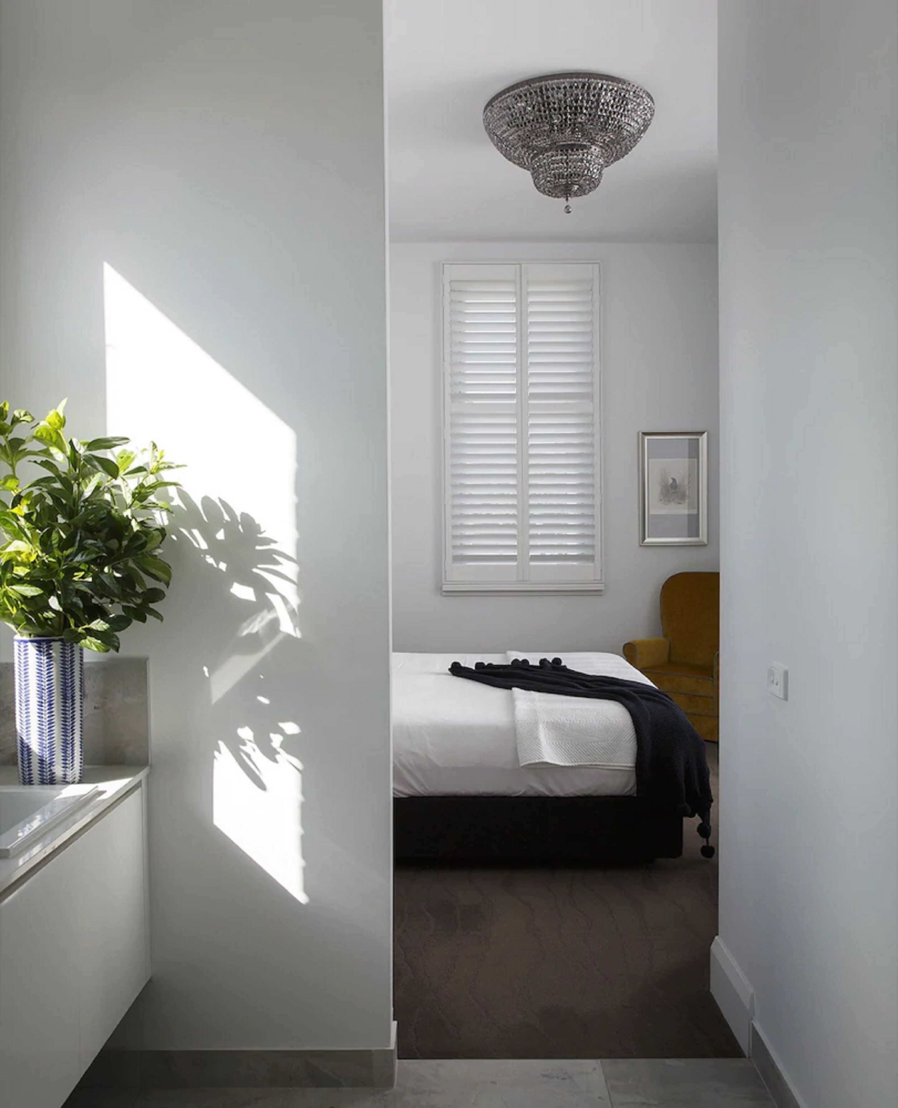 Un pasillo estrecho que conduce a un dormitorio con cama, planta en maceta, ventana con persianas y candelabro.