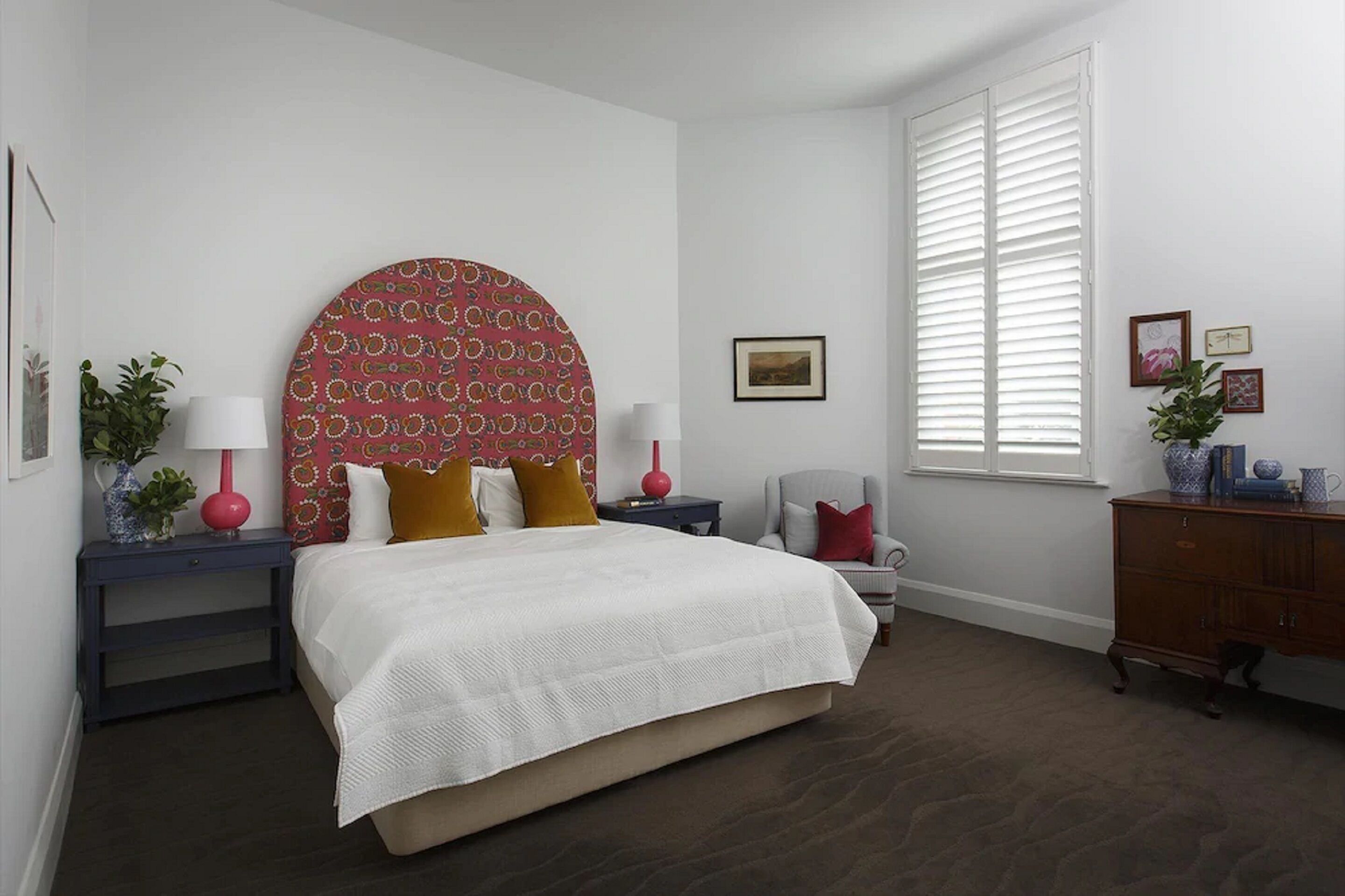 A bedroom with a large bed, a nightstand with a lamp, a chair, a dresser, and a window with blinds.