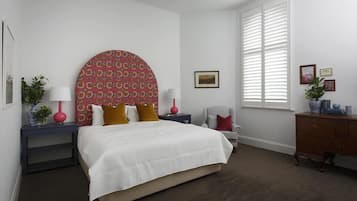 A bedroom with a large bed, a nightstand with a lamp, a chair, a dresser, and a window with blinds.
