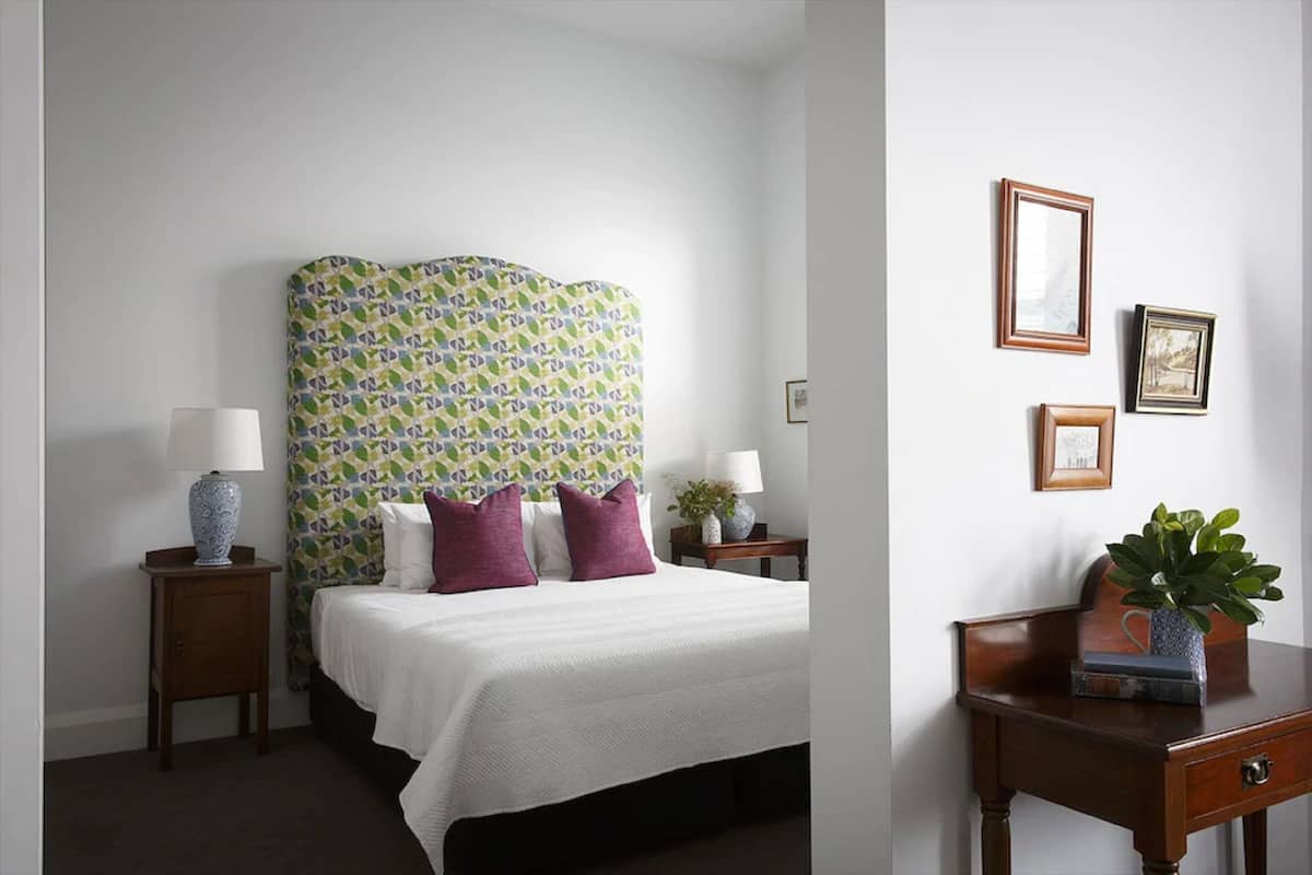 Habitación de hotel con una cama grande, cabecera con estampado llamativo, dos mesitas de noche con lámparas, un escritorio pequeño con una planta y cuadros en la pared.
