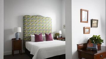 Een hotelkamer met een groot bed, een hoofdbord met een kleurrijk patroon, twee nachtkastjes met lampen, een klein bureau met een plant, en ingelijste foto’s aan de muur.