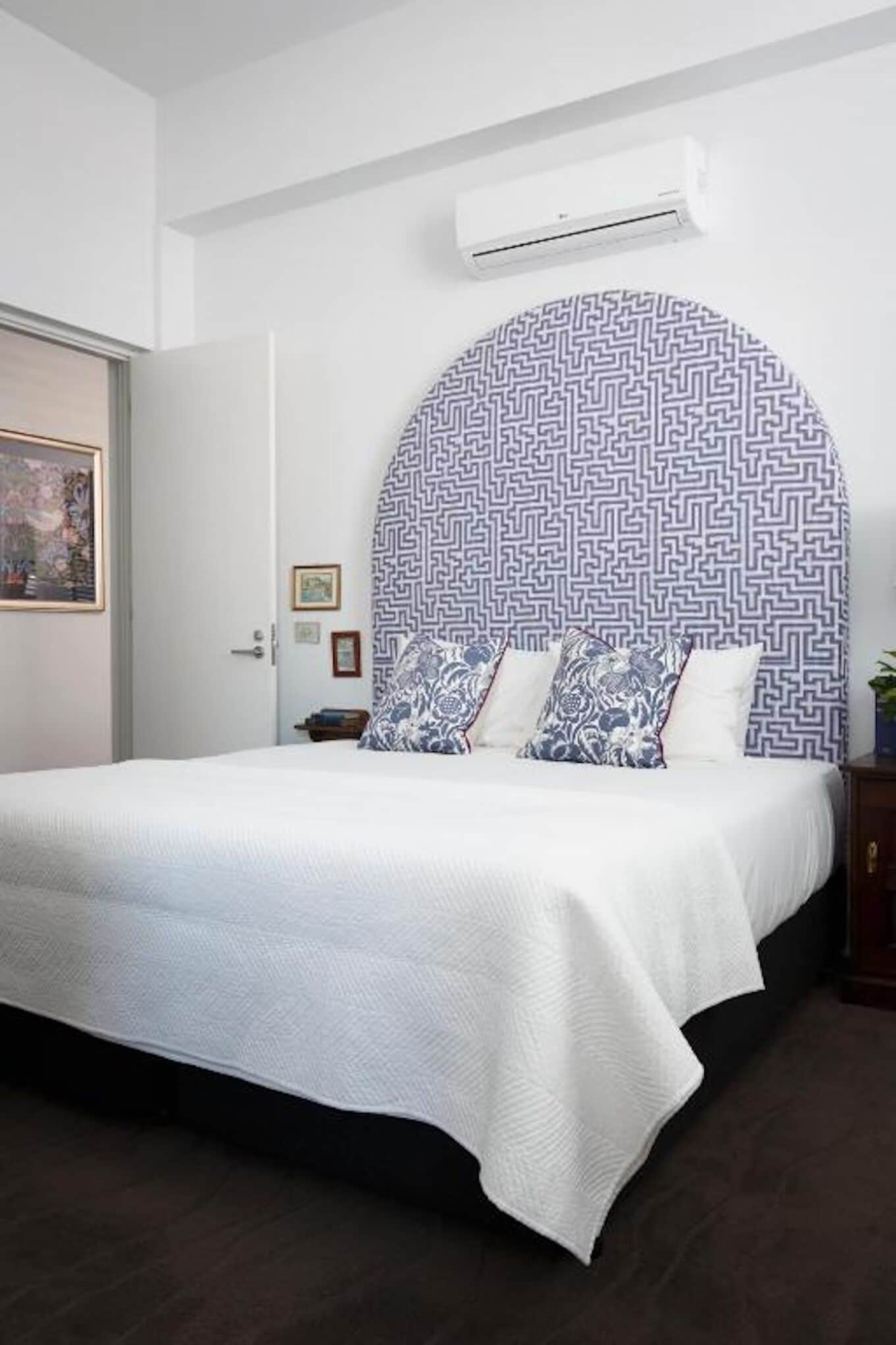 A neatly made bed with a white bedspread and blue patterned pillows, a wall-mounted air conditioner, and a framed picture on the wall.