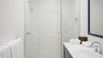 A modern bathroom with a glass-enclosed shower, white tiles, a large mirror, and a white sink with a dark cabinet.