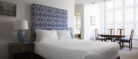 Habitación de hotel impecable con una cama grande, dos mesitas de noche, una mesa de comedor con sillas y un ventanal con persianas.