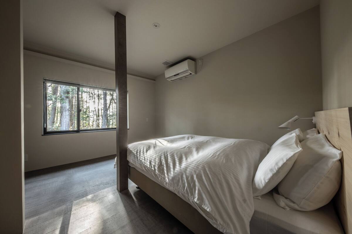 Un dormitorio con cama, ventana con vista a árboles, unidad de aire acondicionado y cabecera de madera.