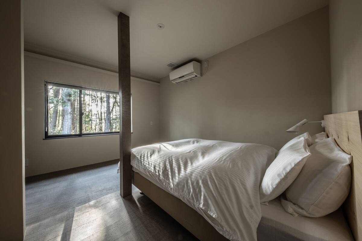 Un dormitorio con un ventanal grande, una cama con ropa blanca y un equipo de aire acondicionado en la pared.