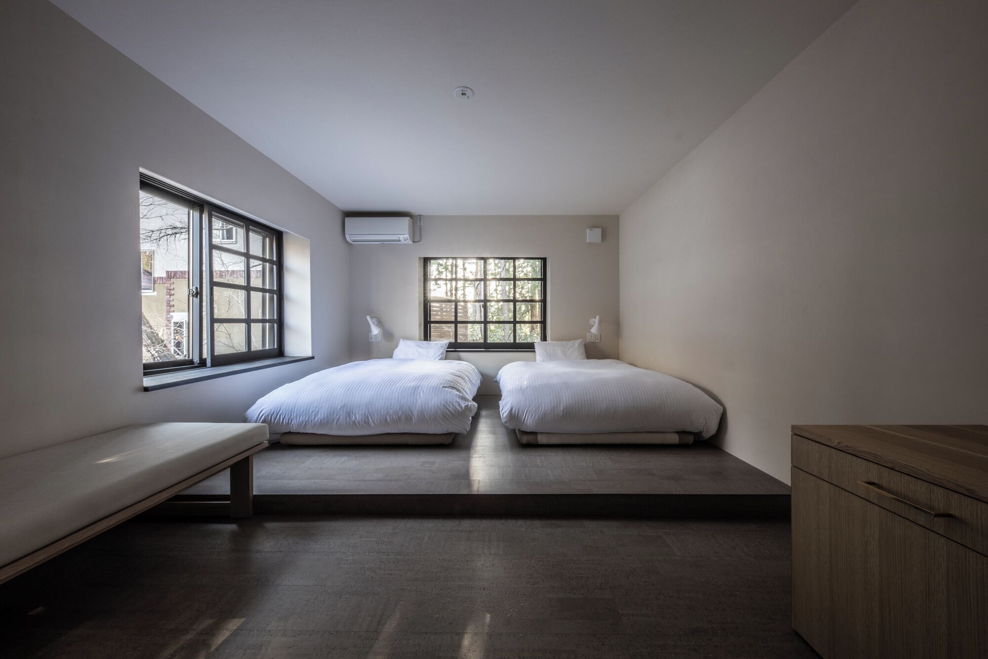 A bedroom with two beds, a bench, a wooden cabinet, and large windows.